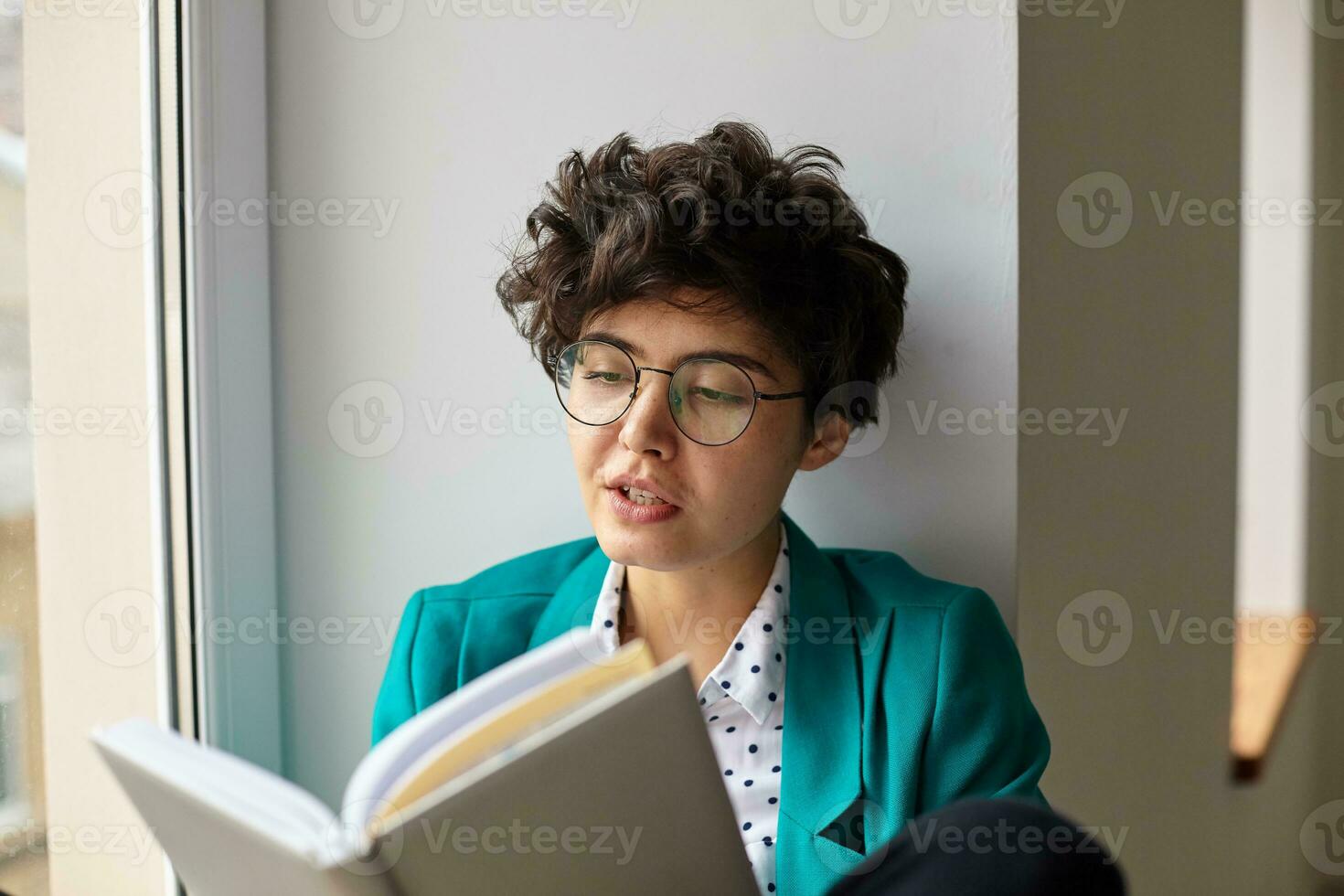 Portrait of charming young curly brunette woman with short trendy haircut keeping book in raised hands while sitting over winowsill on bright day in stylish elegant wear photo