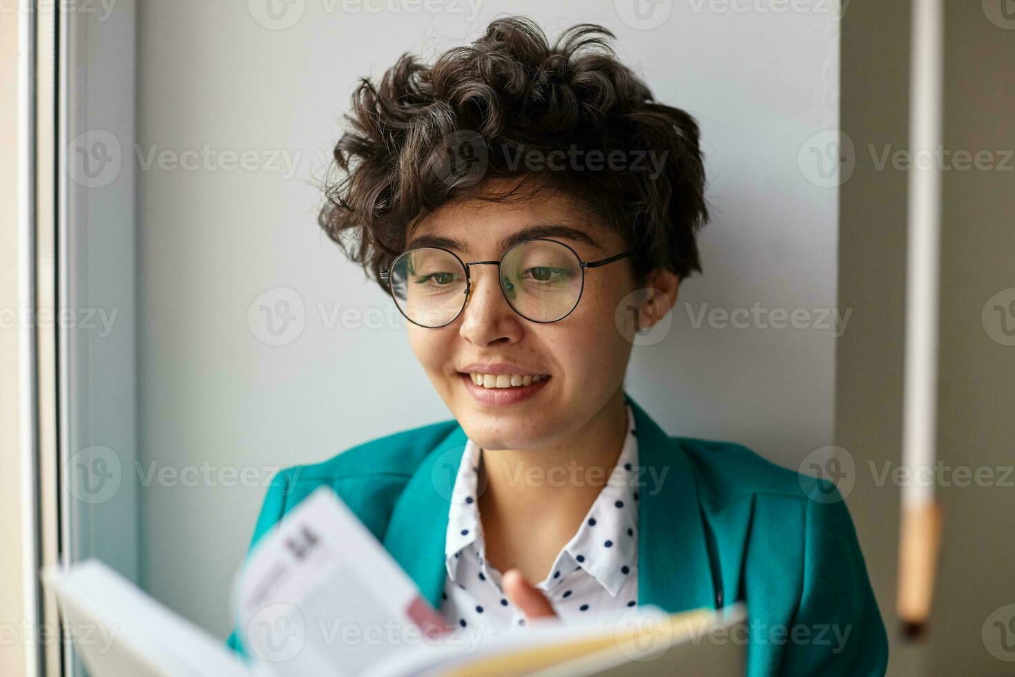 Close-up of positive young brown-eyed short haired curly brunette lady flipping excitedly book and smiling pleasantly while posing over white interior on sunny bright day photo