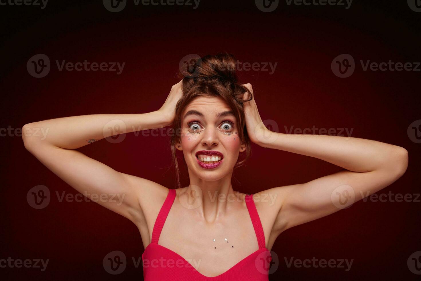 Confused young pretty brunette female with casual hairstyle wearing pink top with straps while posing over burgundy background, clutching her head with pout and rounding eyes wonderingly photo