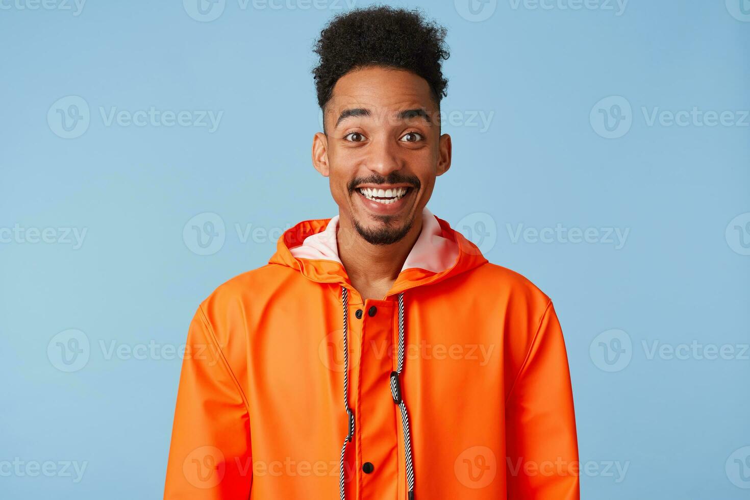 Close up of happy amazed young african american dark skinned man, feels great, wears in orange rain coat, broadly smiles isolated over blue background. photo