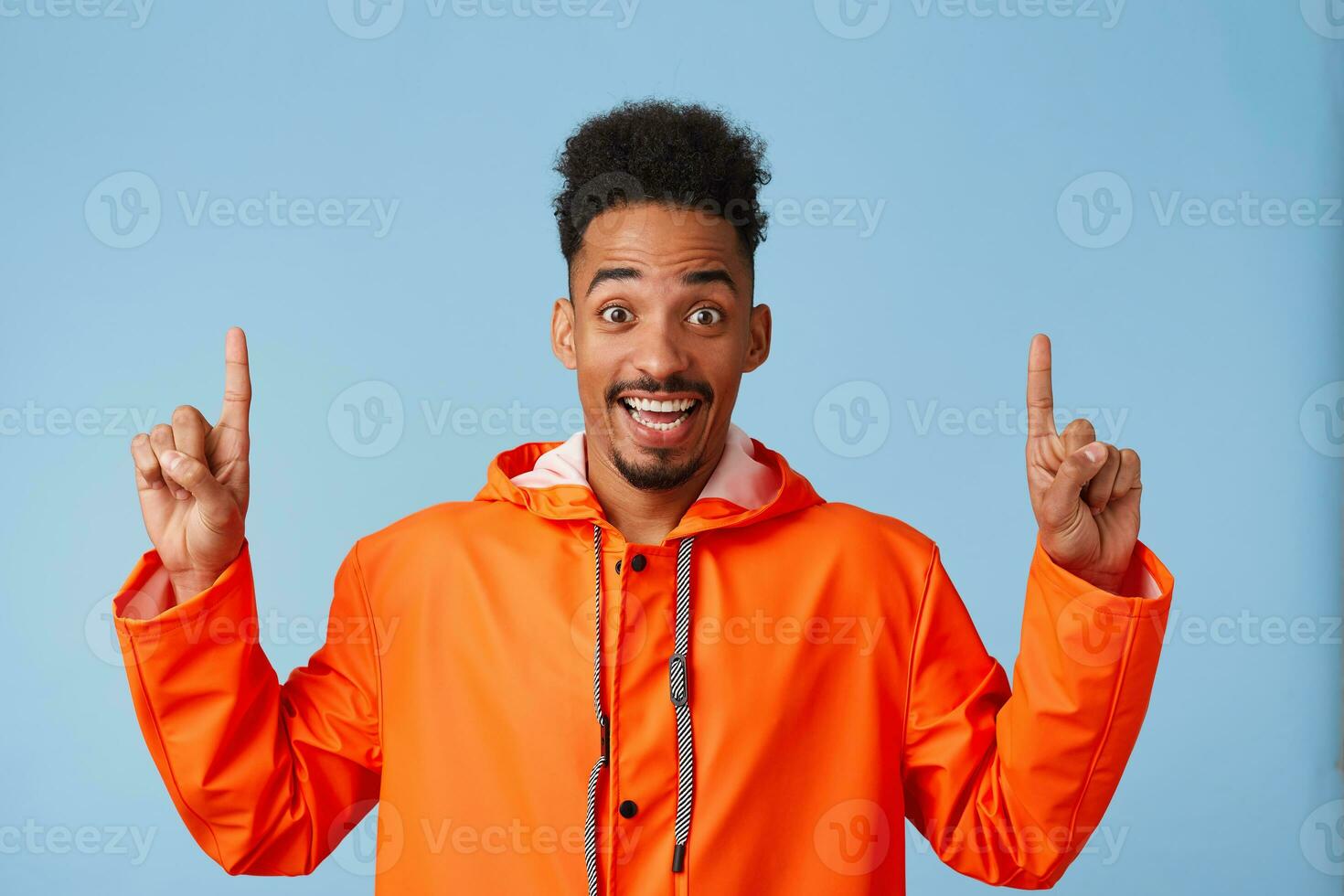 Hey, look up Happy young attractive african american man wears in orange rain coat, smiles broadly stands over blue background and wants to draw your attention at copy space, points fingers up. photo