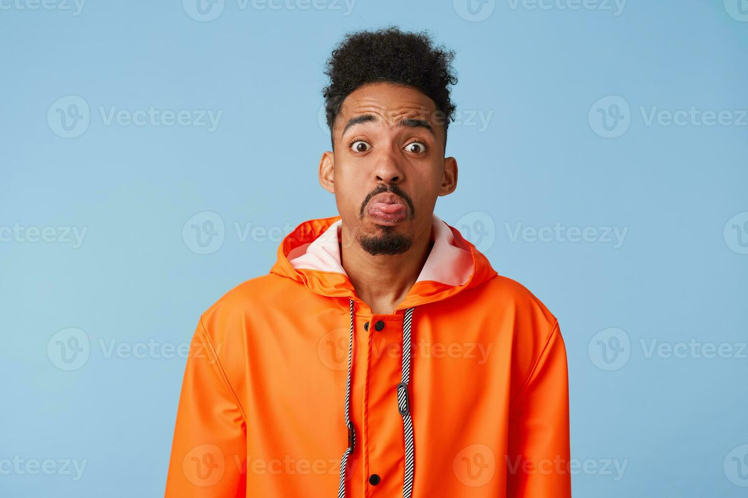 Unhappy african american dark skinned boy wears in orange rain coat,outraged, make grimace, shows tongue stands isolated blue background. photo