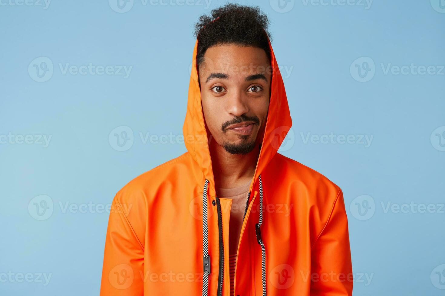 Unhappy handsome african american dark skinned boy wears in orange rain coat, confused, distressed by bad weather and spoiled weekend plans. Looks at the camera over blue background. photo