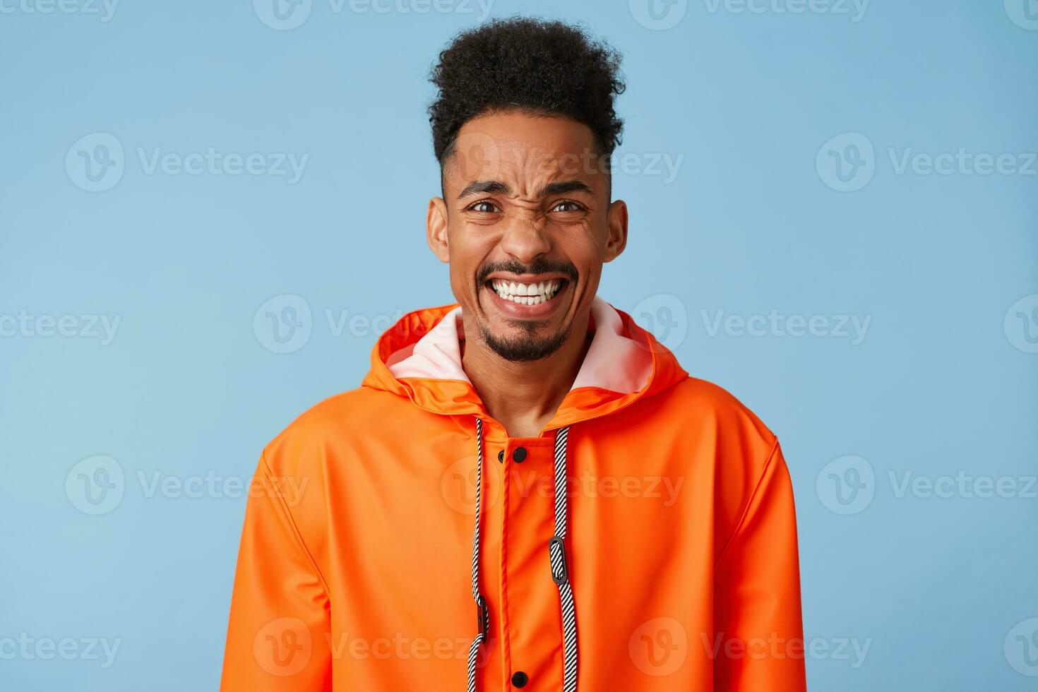 Close up of young attractive african american dark skinned boy wears in orange rain coat, feels very happy and crazy, smiles broadly stands over blue background. photo