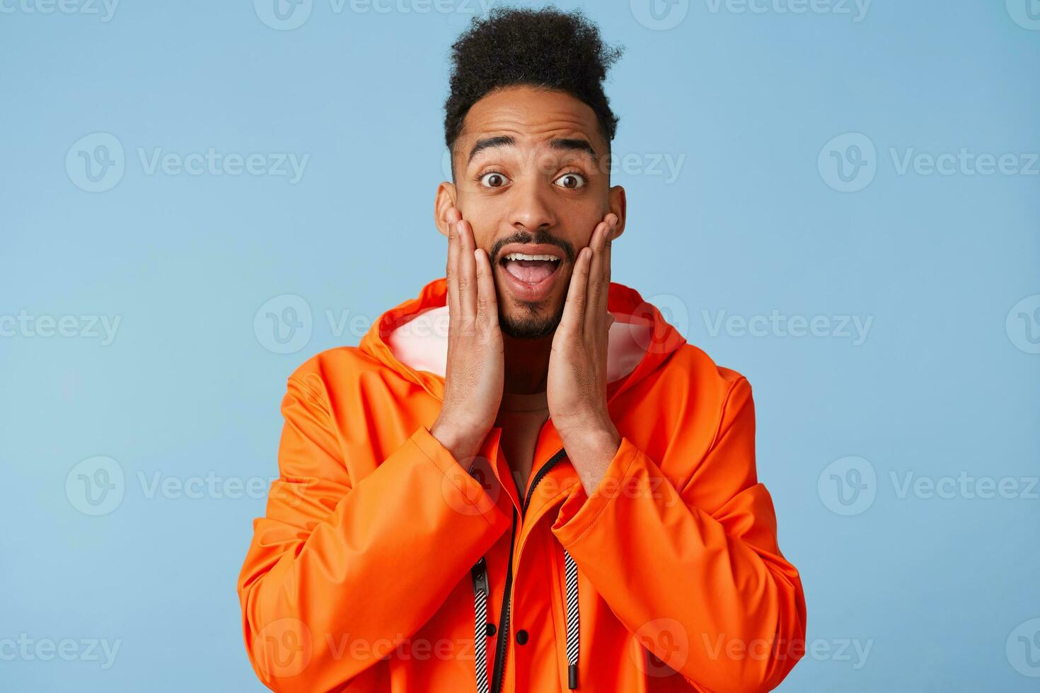 Portrait of shocked young african american dark skinned guy in orange rain coat, touches palms of cheek, can't believe that he saw his idol alive, with wide open mouth, stands over blue background. photo