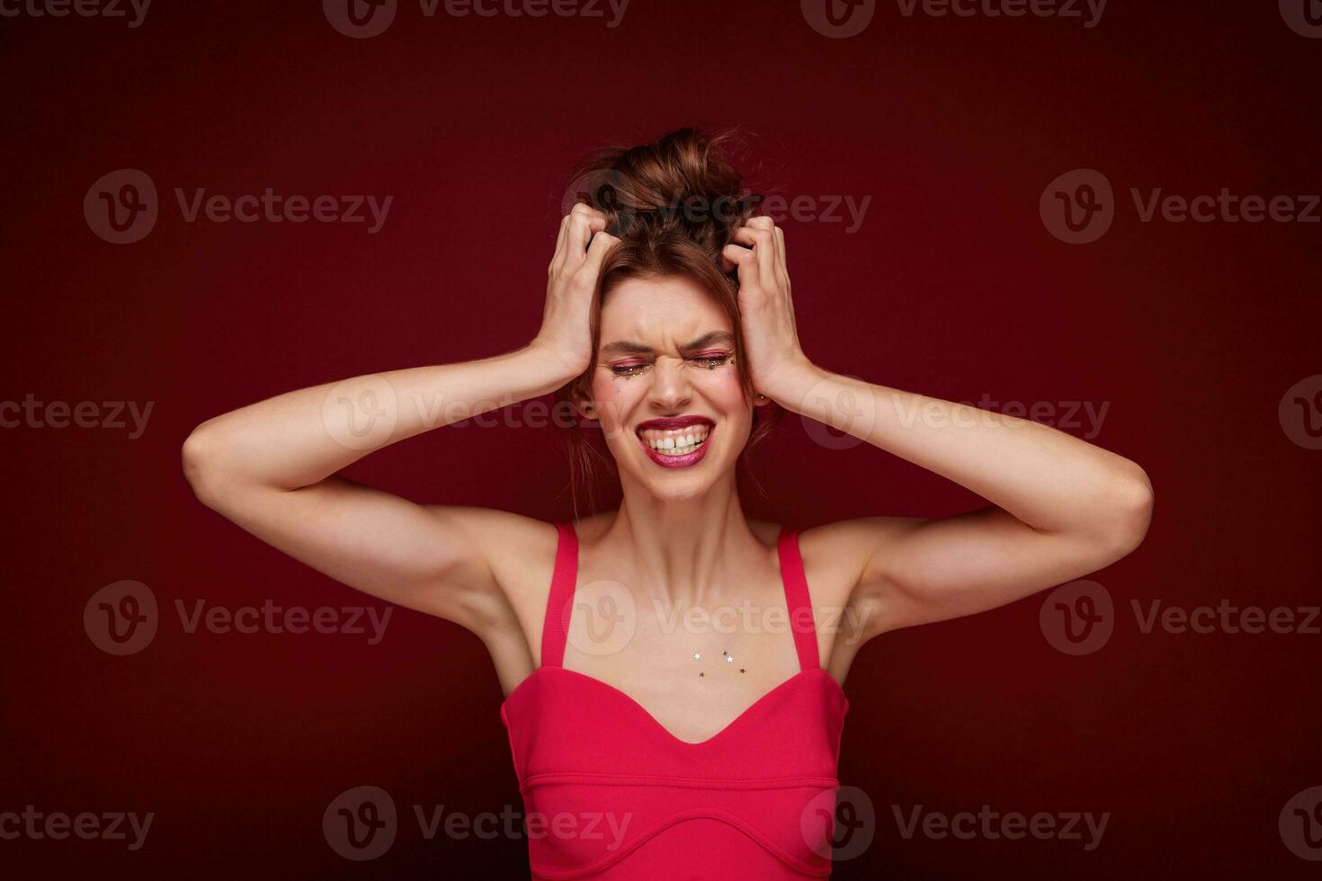 Stressed beautiful young brunette woman with casual hairstyle holding her head with raised hands and frowning discontentedly her face, isolated over burgundy background photo