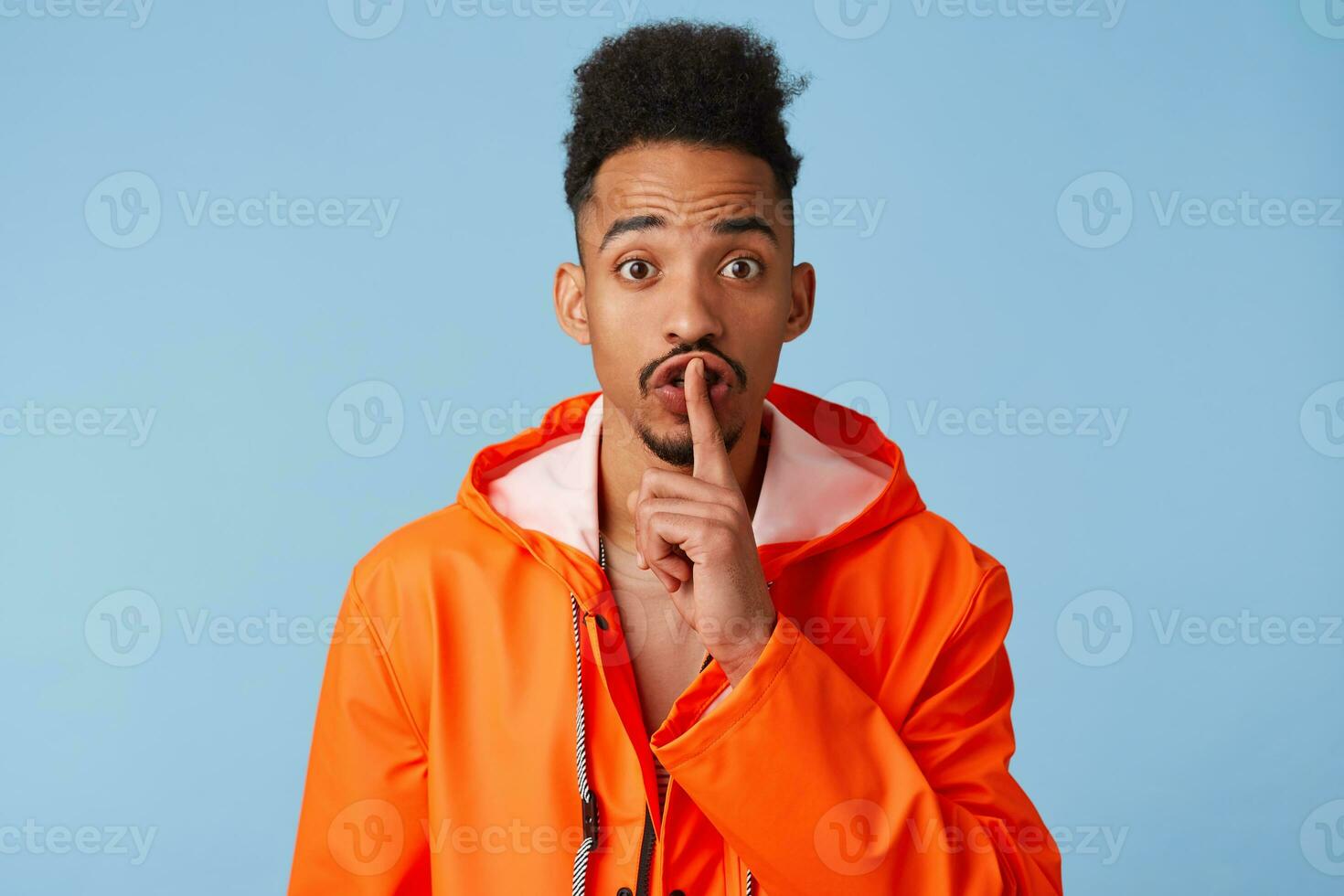 Close up of happy young african american dark skinned man in orange rain coat, tells secret information, demonstrates hush gesture, asks to keep quiet isolated over blue background. photo