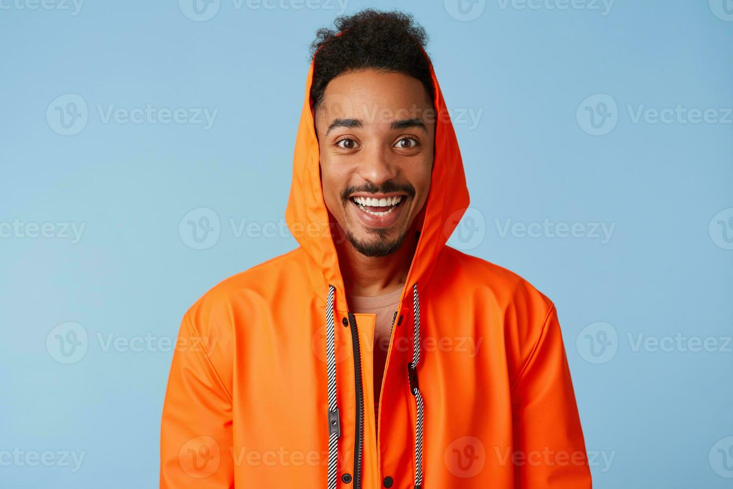 Close up of handsome african american dark skinned boy wears in orange rain coat, feels happy, smiles broadly and stands over blue background. photo