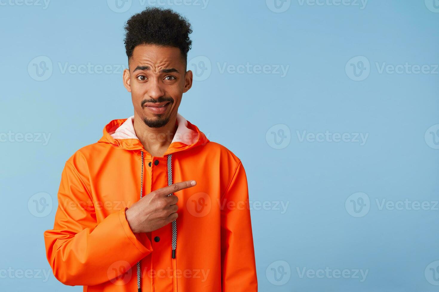 Unhappy young african american guy in orange rain coat looks at the camera inquiringly wants to draw your attention, points fingers at copy space on right. Stands over blue background. photo