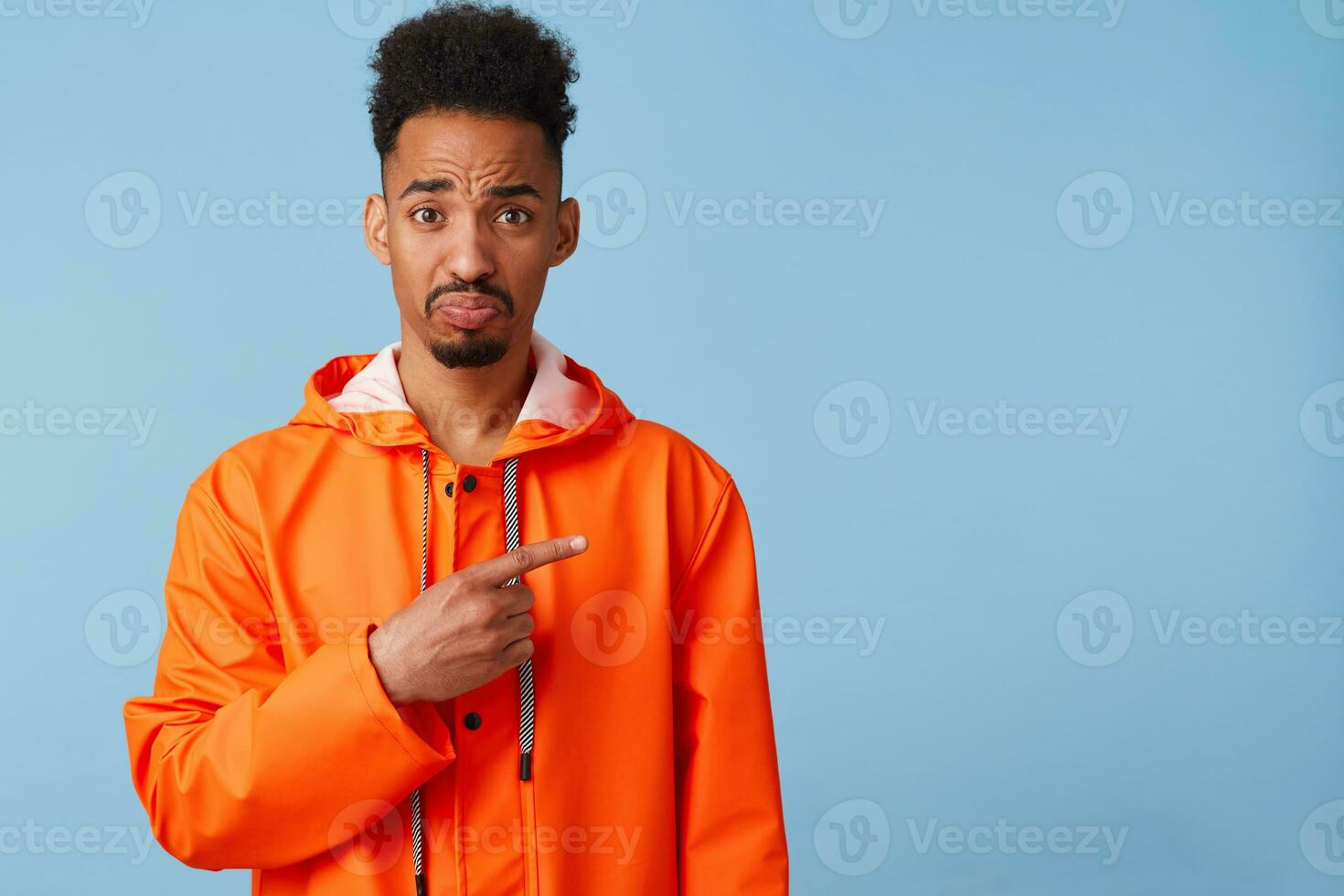 Frustrated young african american guy in orange rain coat, offended looking at the camera, wants to draw your attention, points fingers at copy space on right. Stands over blue background. photo