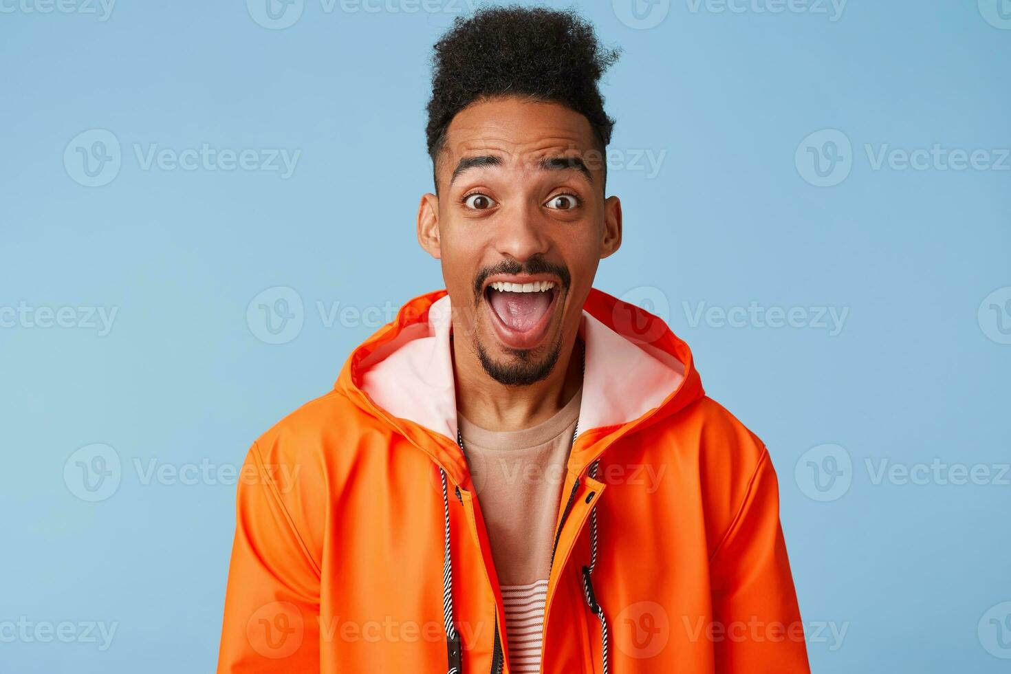 Happy amazed attractive african american dark skinned boy wears in orange rain coat, with wide open mouth, smiles broadly stands over blue background photo