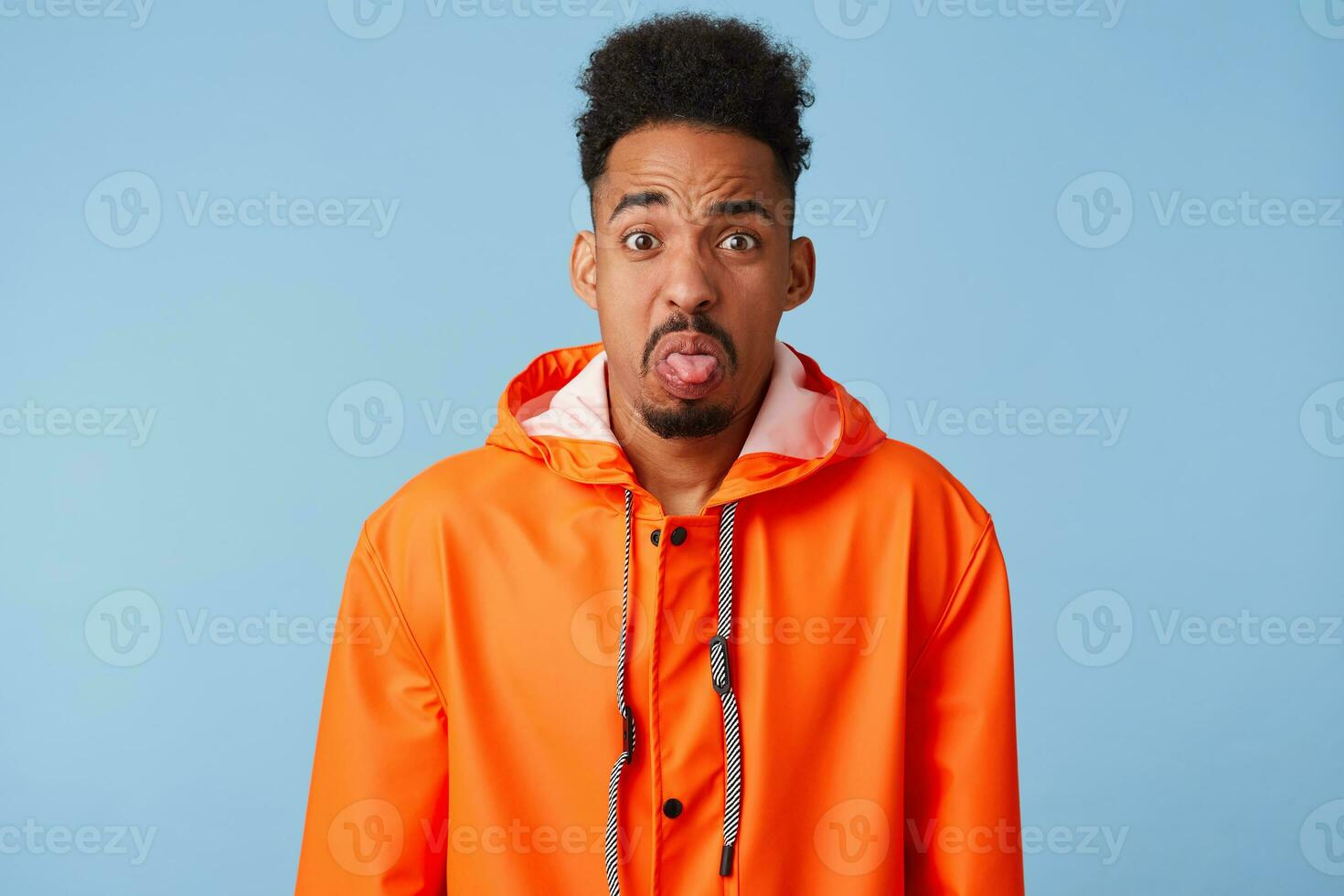 Unhappy african american dark skinned boy wears in orange rain coat,outraged, make grimace, shows tongue as notices something disgusting,stands isolated blue background. photo