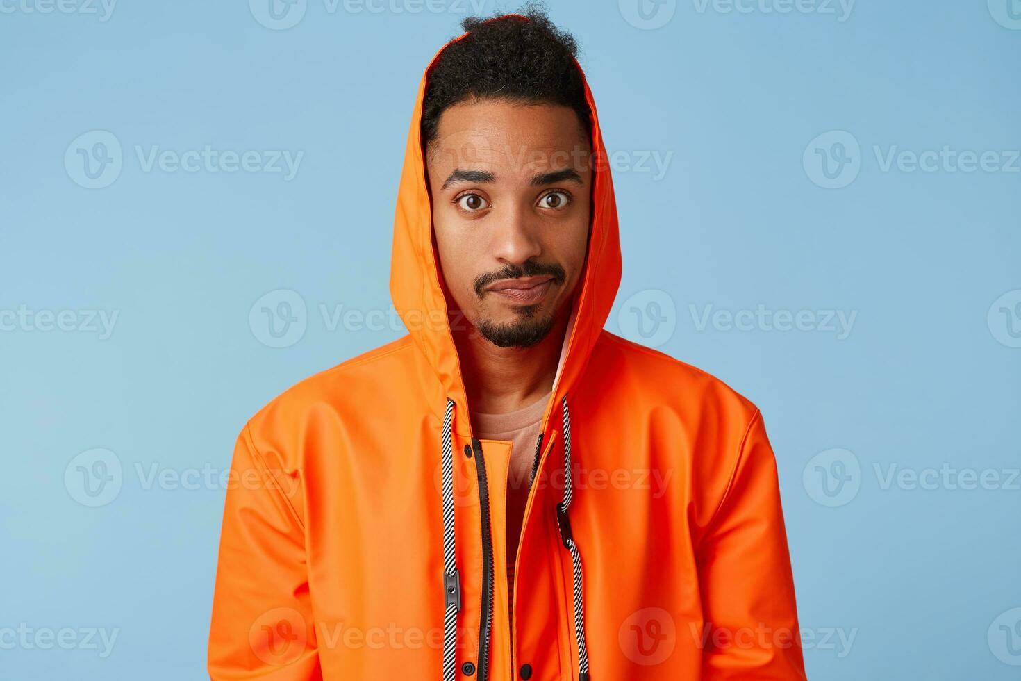 Sad handsome african american dark skinned boy wears in orange rain coat, confused, confused, lost train tickets and can not leave on time. Looks at the camera over blue background. photo