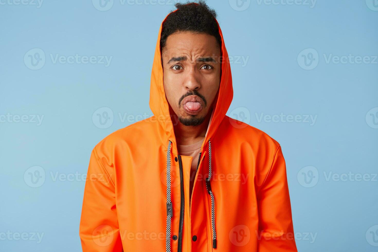 Sad handsome african american boy wears in orange rain coat grimacing, sticking out his tongue at camera, acting like a little naughty child. Looks at the camera over blue background. photo