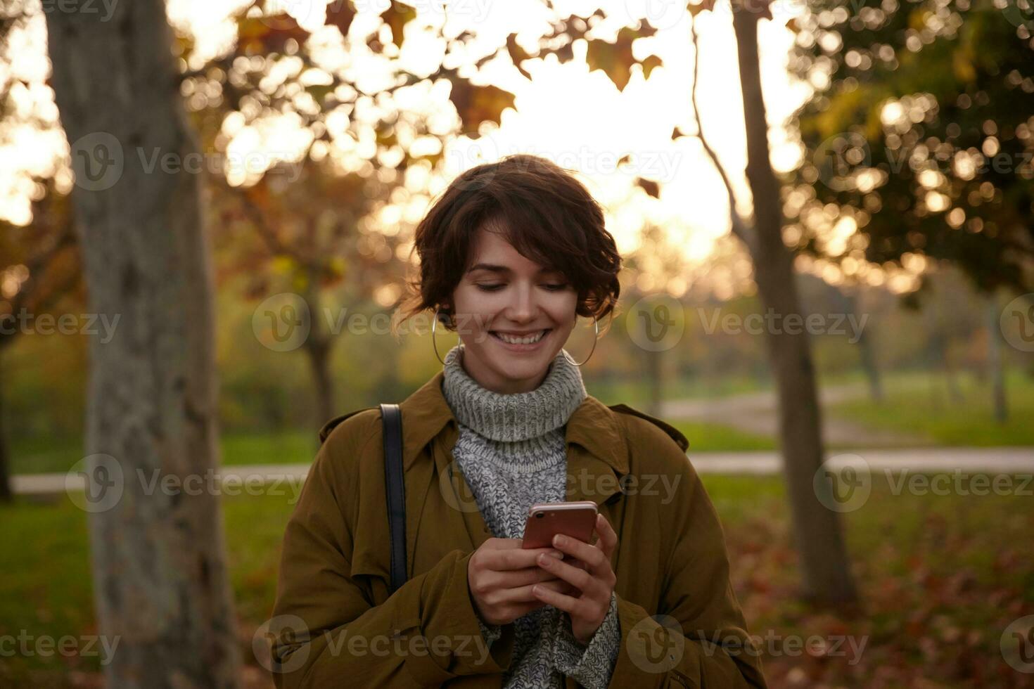 Positive young attractive brown haired woman with natural makeup holding smartphone in raised hands and smiling cheerfully while checking her messages, posing over yellowed trees photo