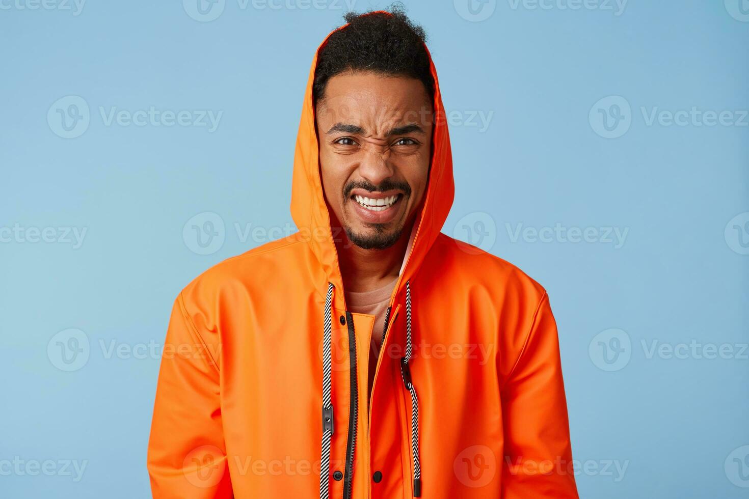 Frowning african american boy wears in orange rain coat grimacing, feel a terrible headache that is unbearable to endure, angry looking at camera standing over blue background. photo