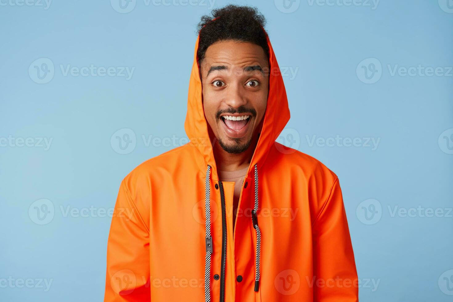 Photo of enthusiastic young african american dark skinned guy in orange rain coat, smiles broadly and looks into the camera with wide open mouth, stands over blue background.