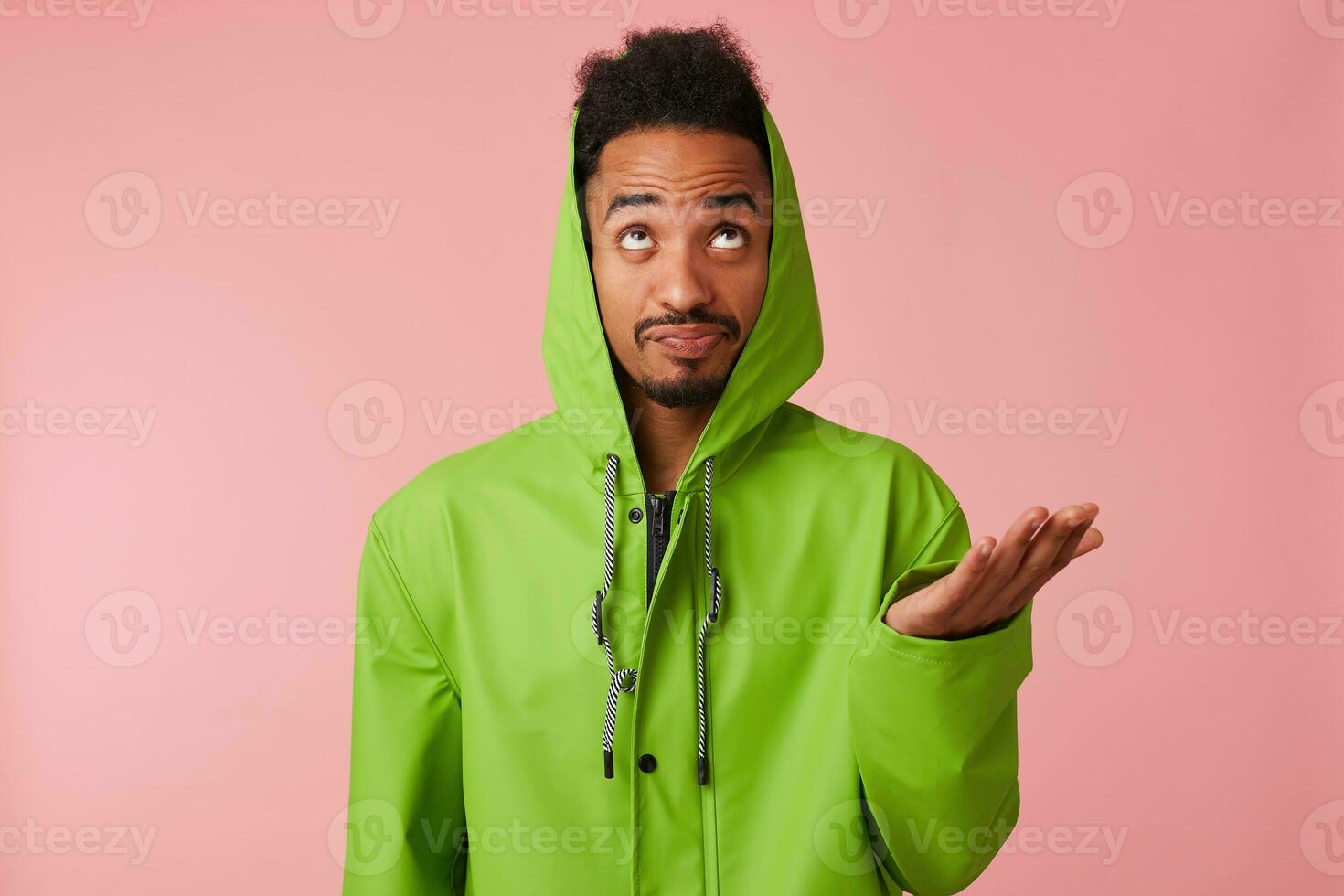 Close up of displeased young African American handsome guy in green rain coat, querulous looks up at the copy space, stands over pink background. photo