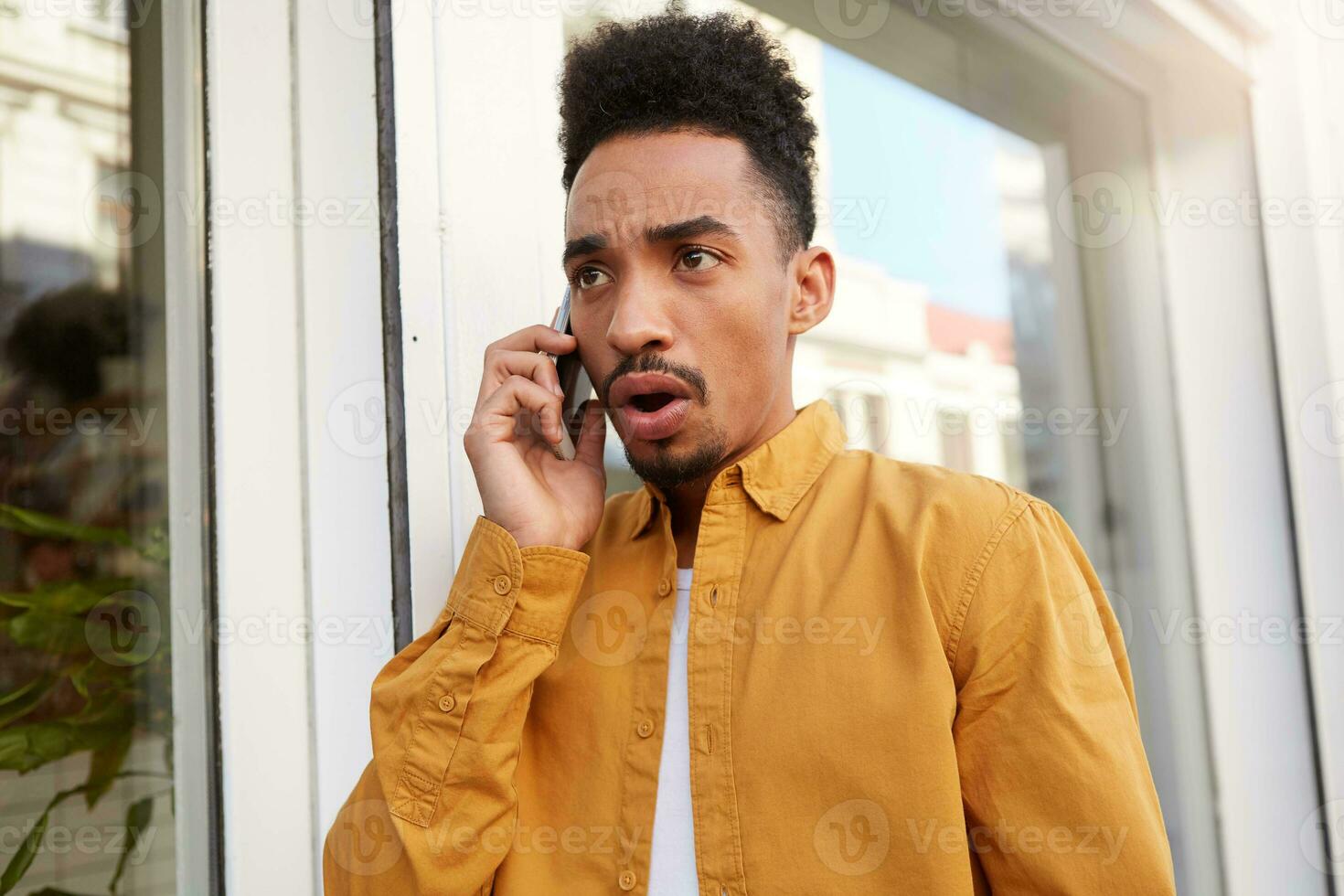 joven oscuro desollado asombrado oscuro desollado hombre en amarillo camisa, habla en el teléfono con su amigos y caminando abajo el calle, con conmocionado expresión. foto