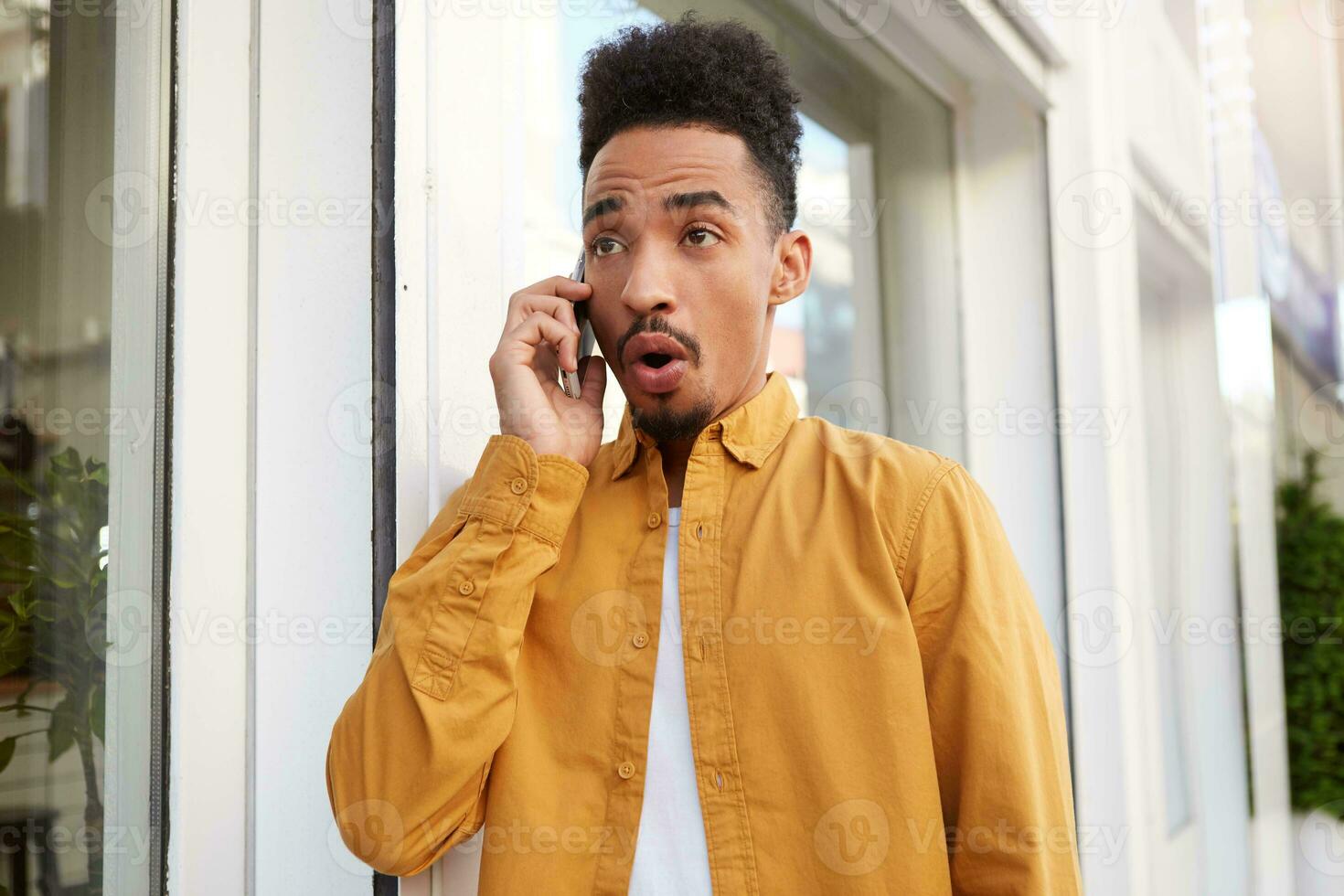 Portrait of young shocked dark skinned man in yellow shirt, walking down the street and speaks on the phone with his friends, hears unbelievable news, wit wide open mouth and eyes. photo