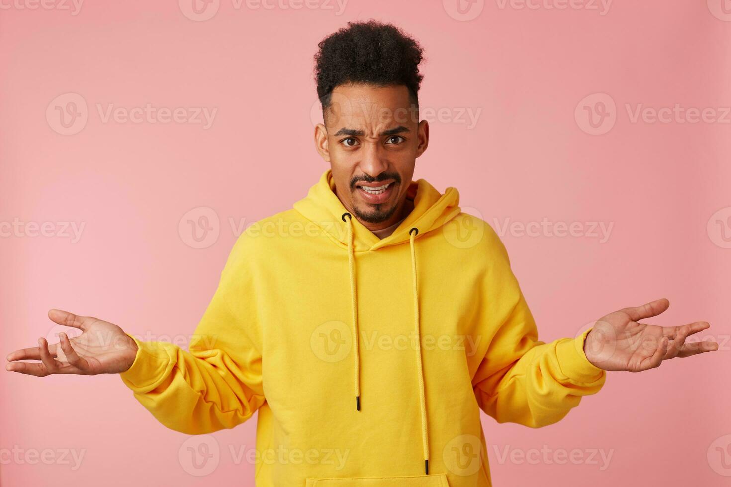 Photo of young displeased african american guy in yellow hoodie, frowning and throws up his hands in a misunderstanding of what is happening, looking at the camera over pink background.