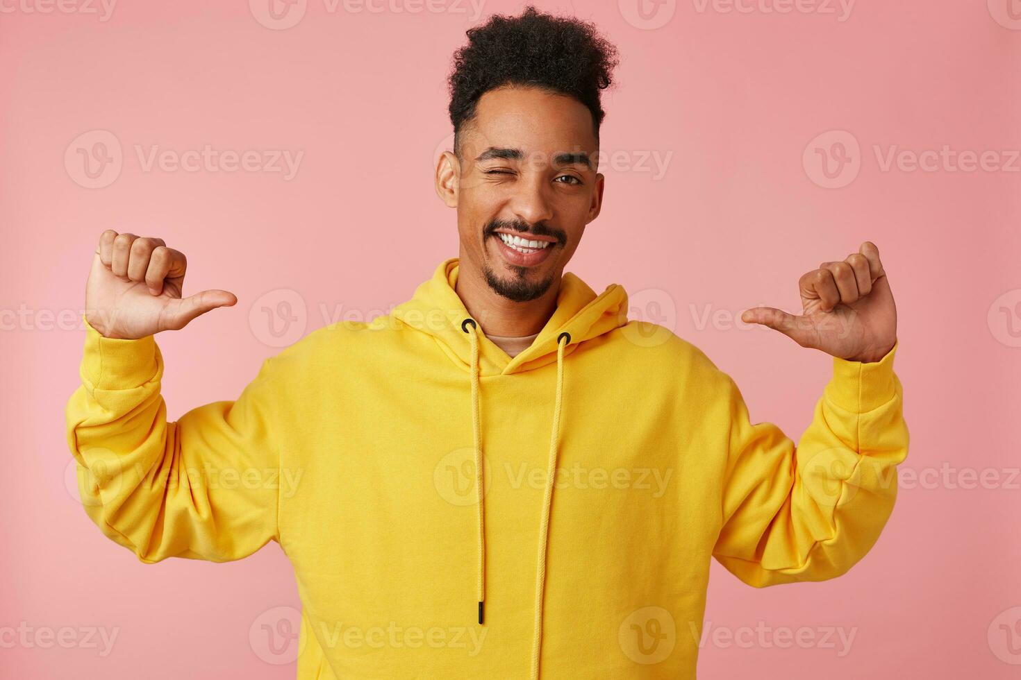joven alegre africano americano chico en amarillo sudadera, levantamiento su manos puntos a él mismo con su pulgares, decir soy Frío' y soportes terminado rosado antecedentes con Copiar espacio. foto