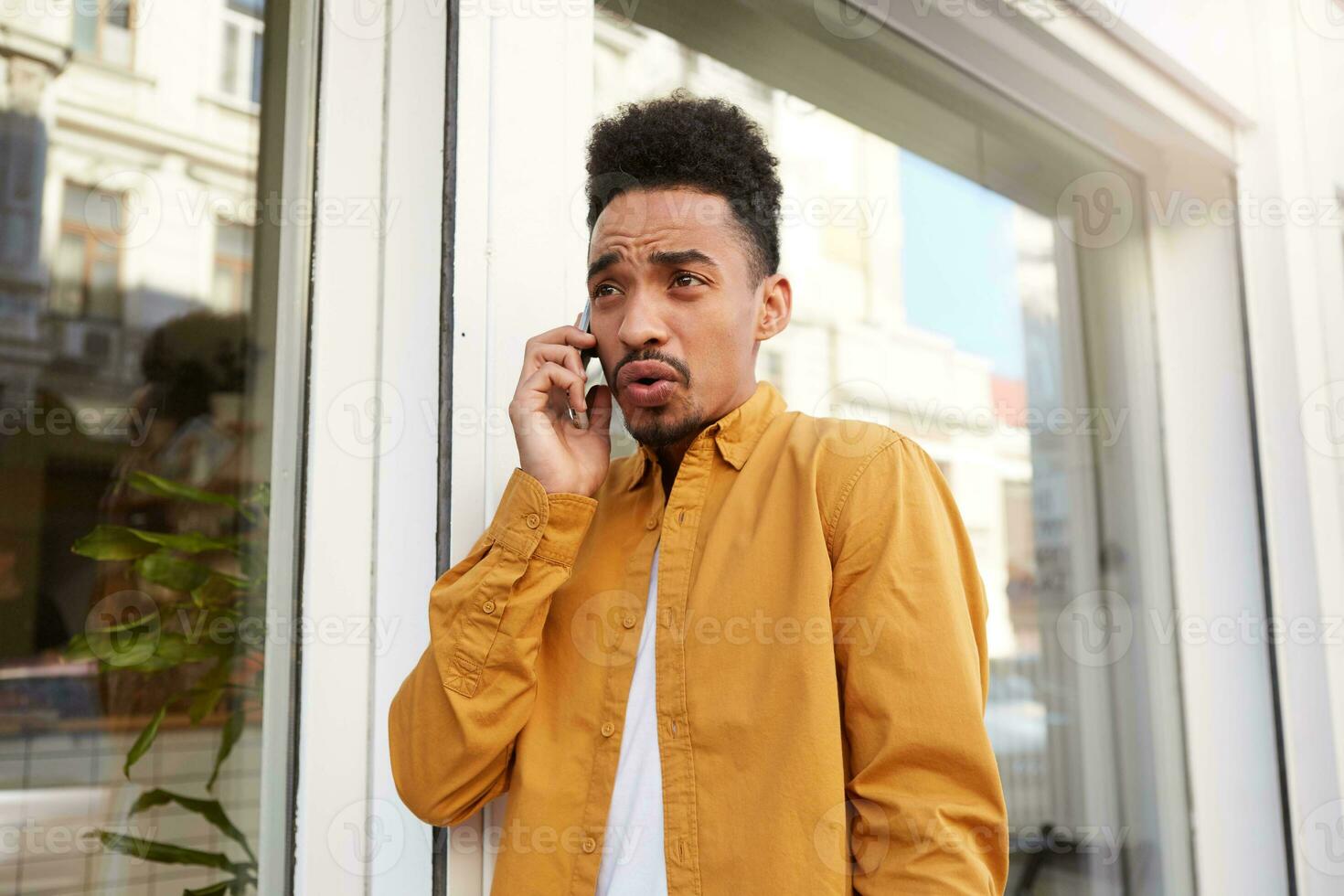 joven insatisfecho oscuro desollado hombre en amarillo camisa, habla en el teléfono con su amigos y caminando abajo el calle, con resentido expresión. foto