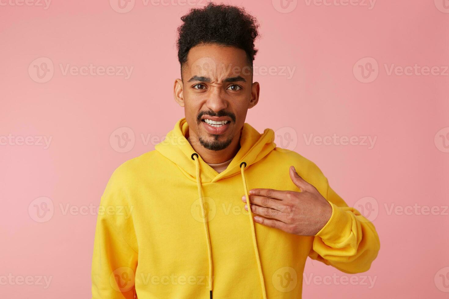 Portrait of young displeased african american guy in yellow hoodie, points to himself in a misunderstanding of what is happening, looking at the camera over pink background. photo