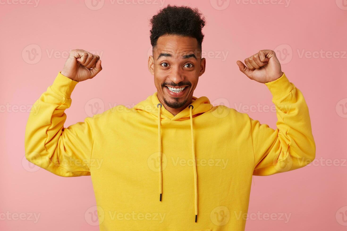 joven alegre africano americano hombre en amarillo sudadera, levantamiento su puños arriba, decir soy Frío' y soportes terminado rosado antecedentes y sonriente en general. foto