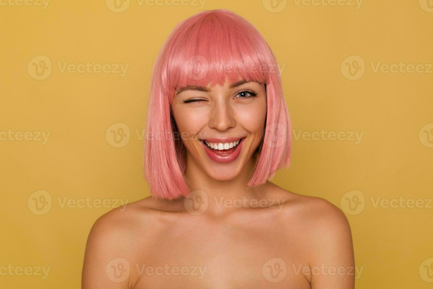 Joyful young attractive female with short pink hair keeping one eye closed while looking cheerfully at camera, being in nice mood and smiling broadly, isolated over mustard background photo