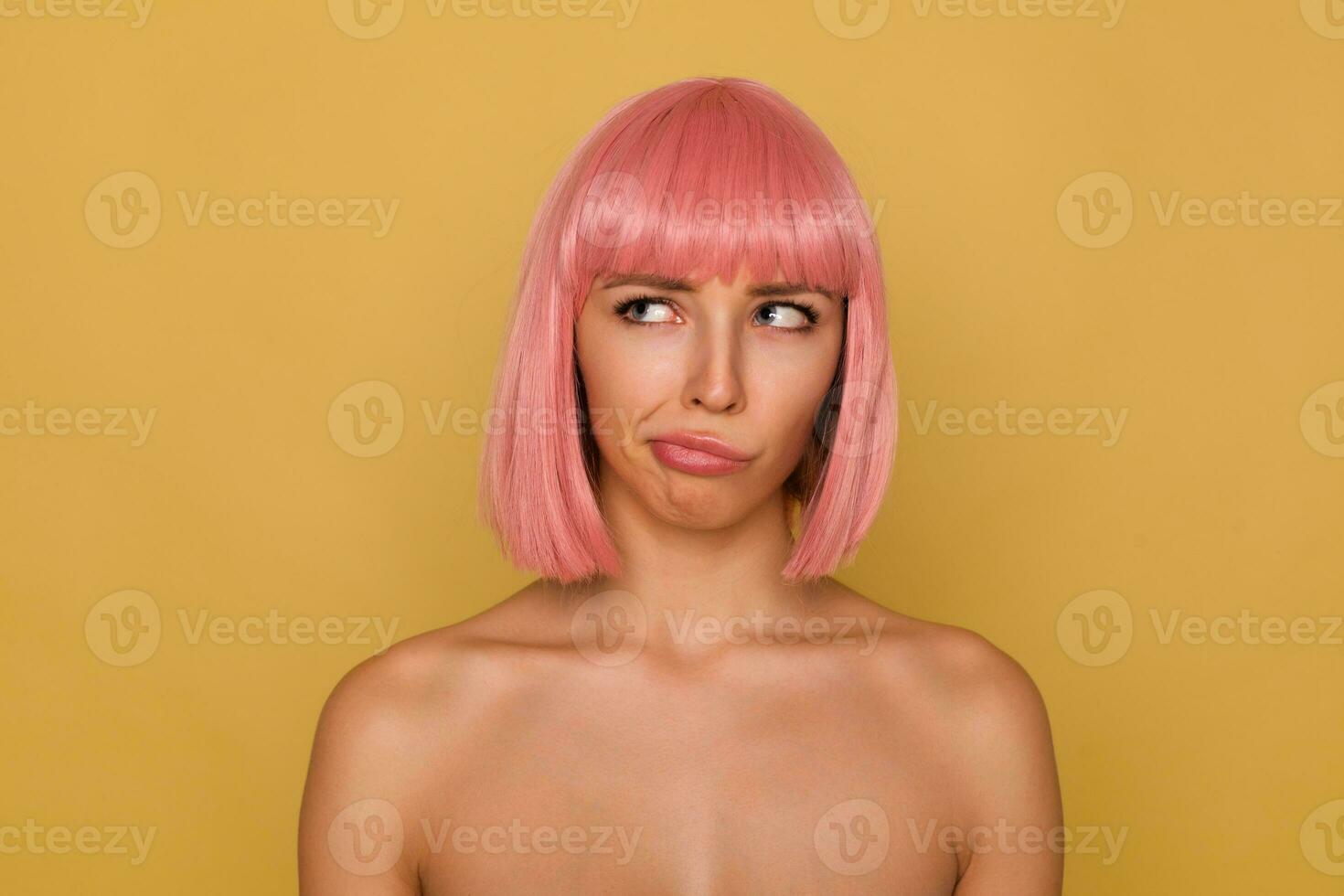 perplejo joven atractivo rosado peludo hembra con natural maquillaje retortijón su boca mientras mirando confusamente hacia arriba, posando terminado mostaza antecedentes con desnudo espalda foto