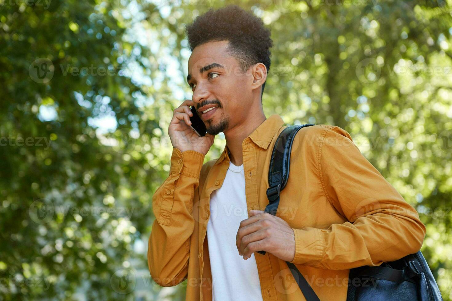 Young positive African American guy walking at the park and speaking on telephone, wears in yellow shirt, waitings his friends and looks away. photo