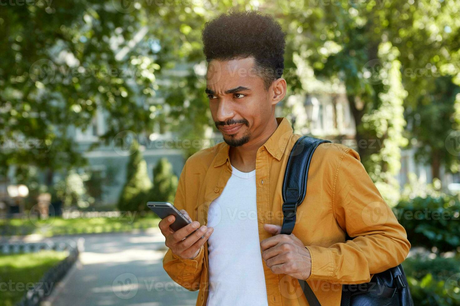 Young dissatisfied dark skinned guy, chatting on the phone with his friends and walking at the park, looking from under his forehead with resentful expression, his girlfriend is late again. photo