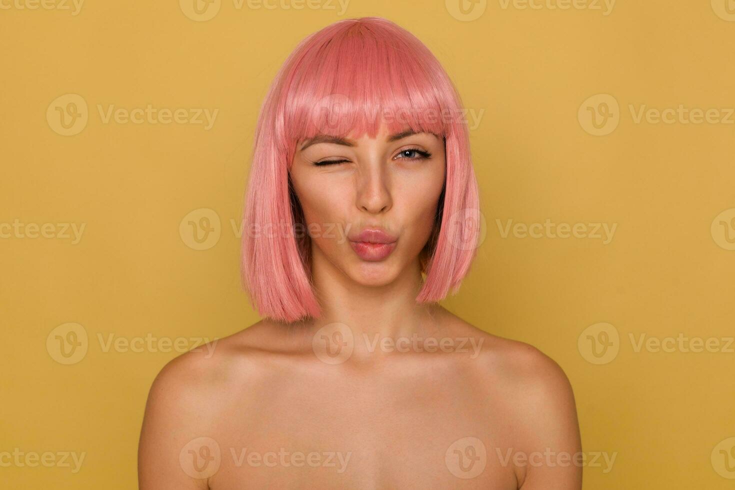 Portrait of flirty pretty young woman with pink bob haircut pouting her lips in air kiss and giving wink while looking positively at camera, posing over mustard background photo