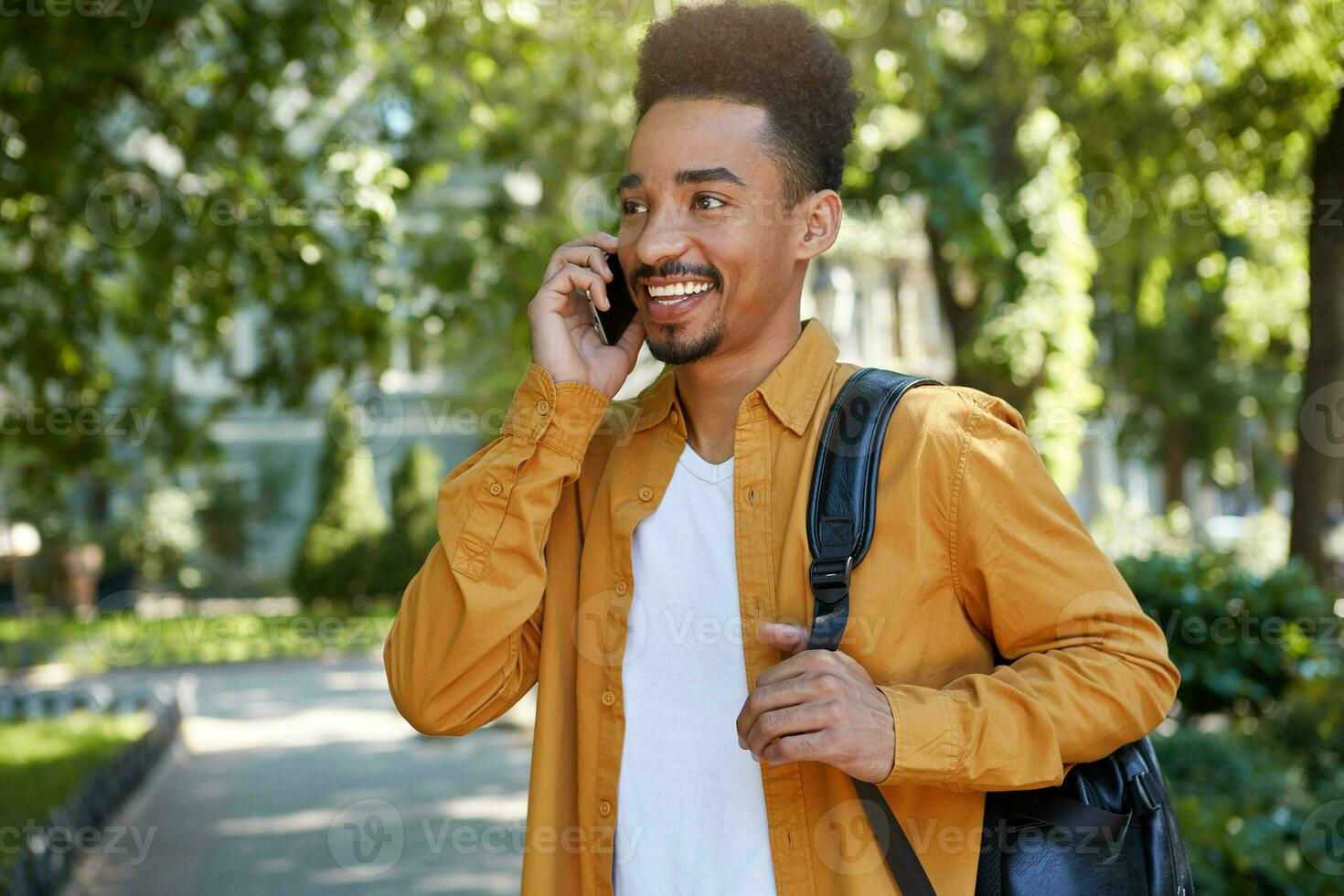 Young smiling dark skinned student walking at the park, speaking on smartphone, waitng his friend, wears in yellow shirt, looking away and broadly smiling. photo