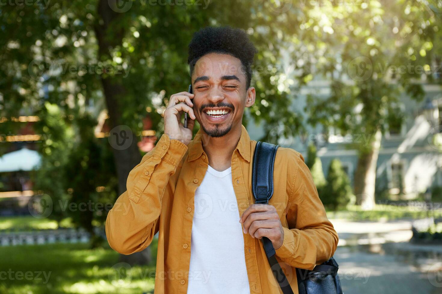 Portrait of young dark skinned smiling man walking in the park in a yellow shirt and a white T-shirt, with a backpack on one shoulder, talking on the phone with his friend, smiling and enjoys walk. photo