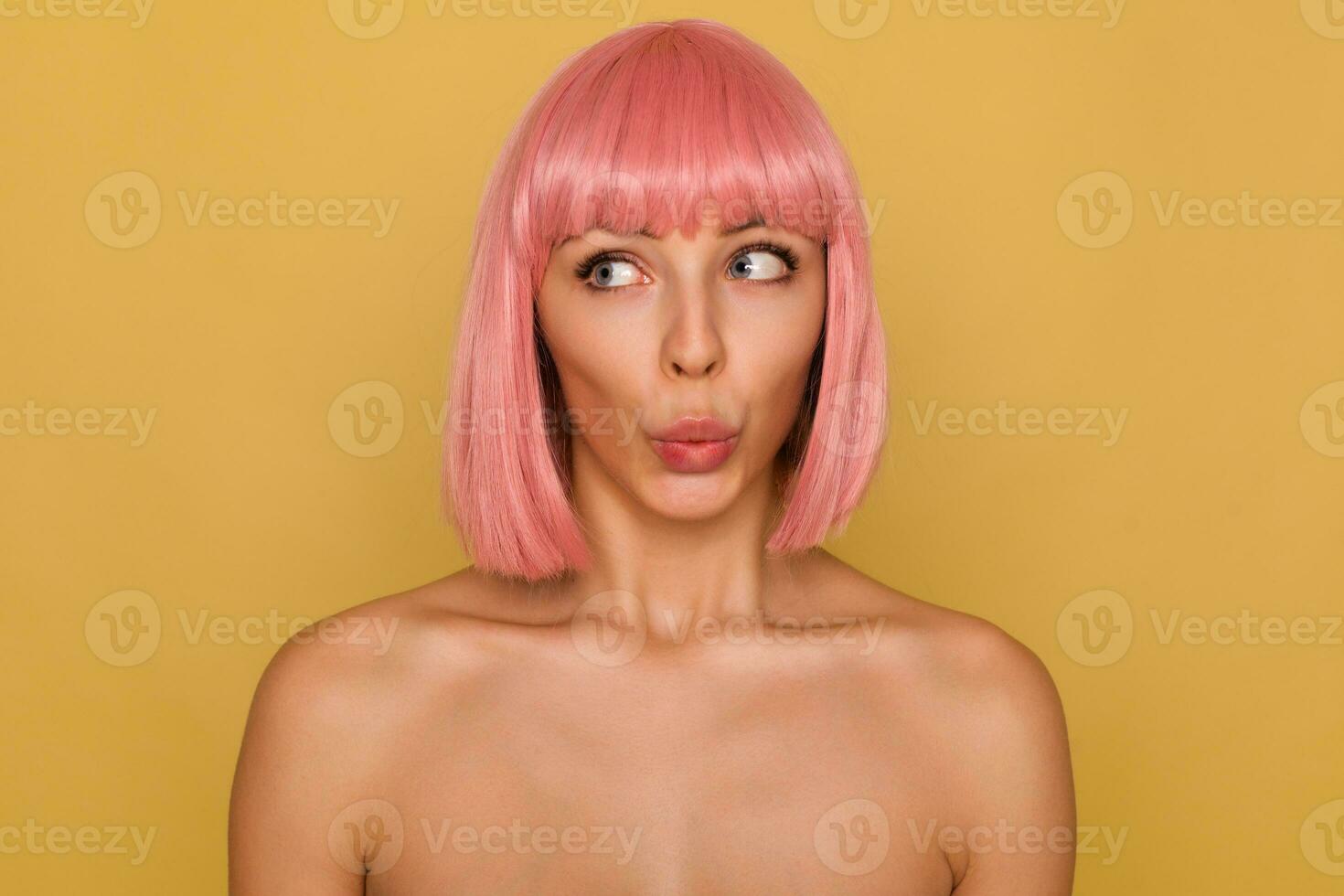 Close-up of excited young beautiful lady with short pink hair folding her lips while looking confusedly aside, posing over mustard background with naked shoulders photo