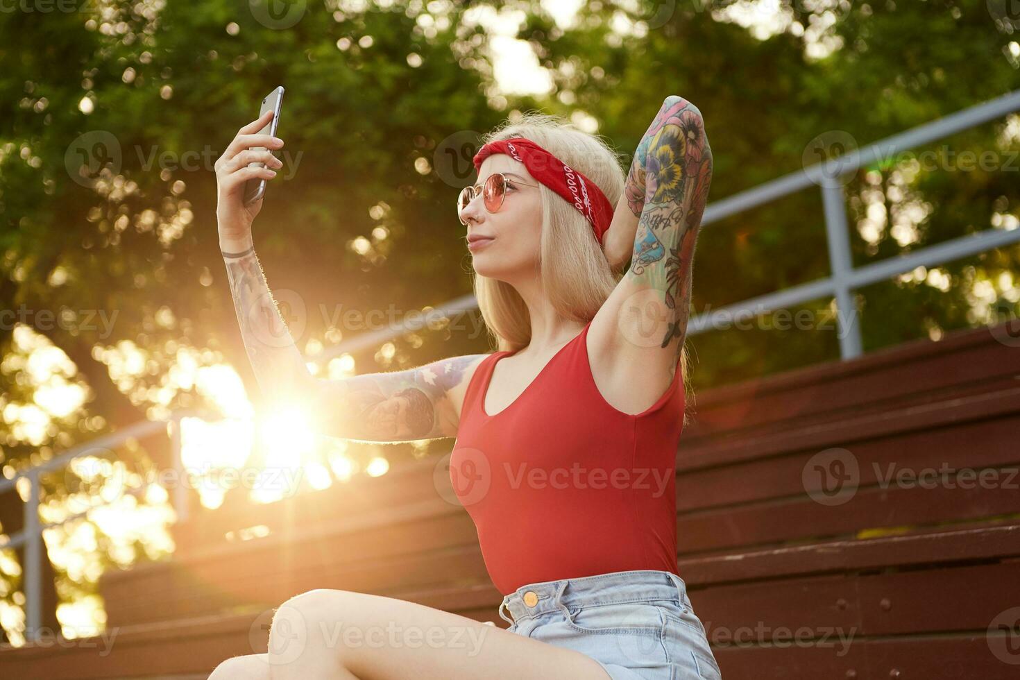 retrato de joven hermosa rubia mujer con tatuado brazos participación teléfono inteligente y hace selfie para su Blog. usa en un rojo camiseta y mezclilla bermudas, con un de punto pañuelo en cabeza, en rojo lentes. foto