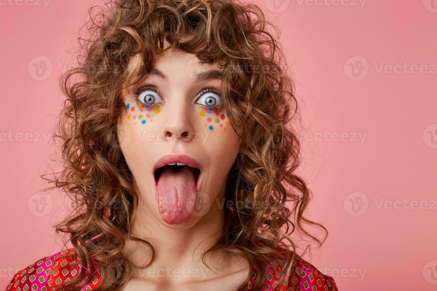 Poratrait of young curly blue-eyed woman with multicolored dots on her face looking at camera with wide eyes opened and sticking out her tongue, isolated over pink background photo