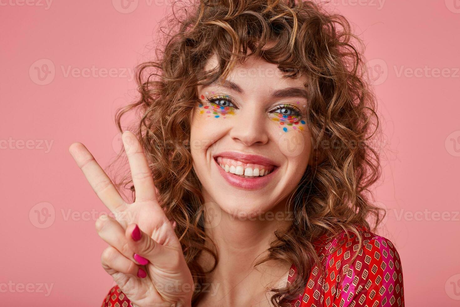 curly blue-eyed female with festive hairstyle and multicolored dots on her face raising hand with victory sign, looking joyfully to camera and smiling widely, posing over pink background photo