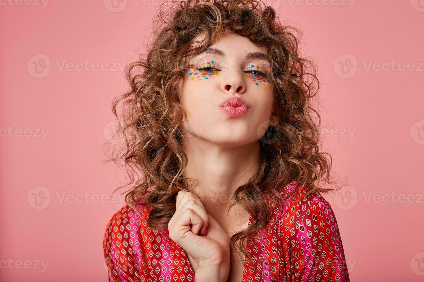 Cheerful young curly brunette female with festive hairstyle and multicolored dots on her face standing over pink background, folding lips in air kiss and looking joyfully to camera photo