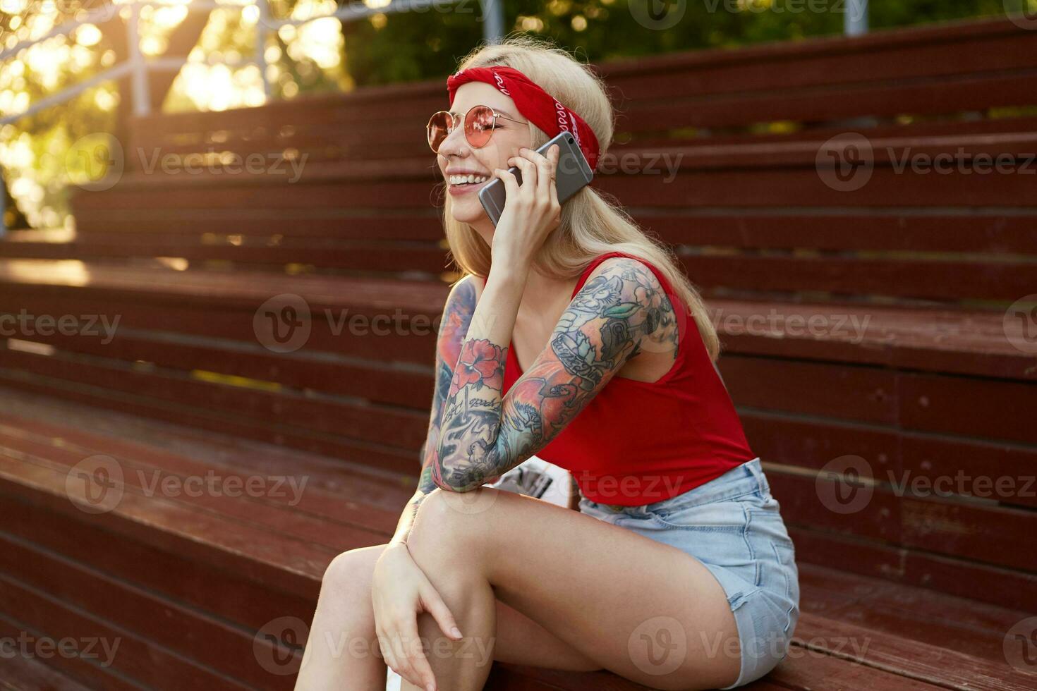 Portrait of young nice blonde woman with tattooed arms, holding a smartphone at the ear and talking to his friend. ears in red T-shirt, denim shorts, with knitted bandana on her head, in red glasses. photo