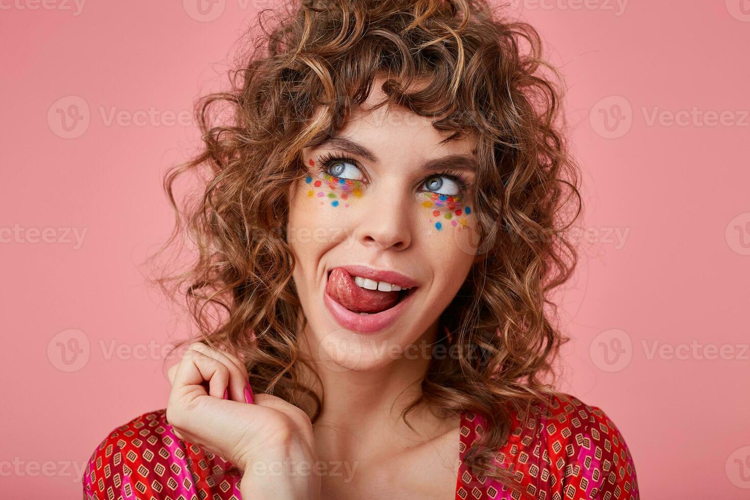Studio photo of pretty young blue-eyed lady with brown curly hair and festive makeup posing over pink background in colored petterned top, looking aside cheerfully and showing her tongue