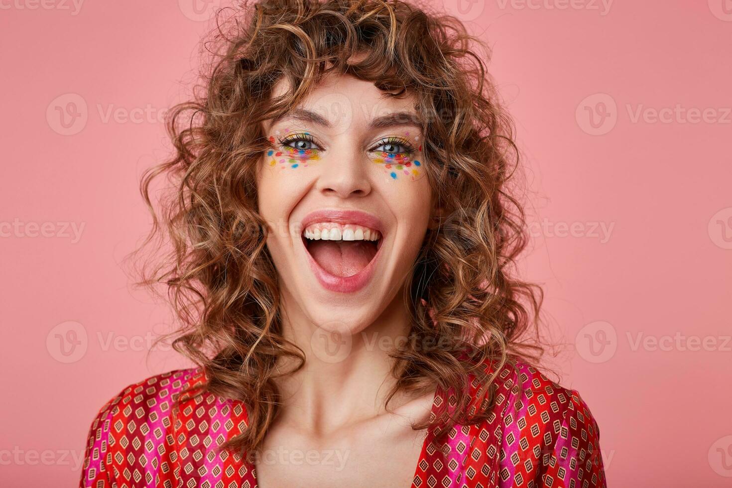 Overjoyed young blue-eyed woman with brown curly hair looking at camera happily and laughing with wide mouth opened, isolated over pink background in colored patterned top photo