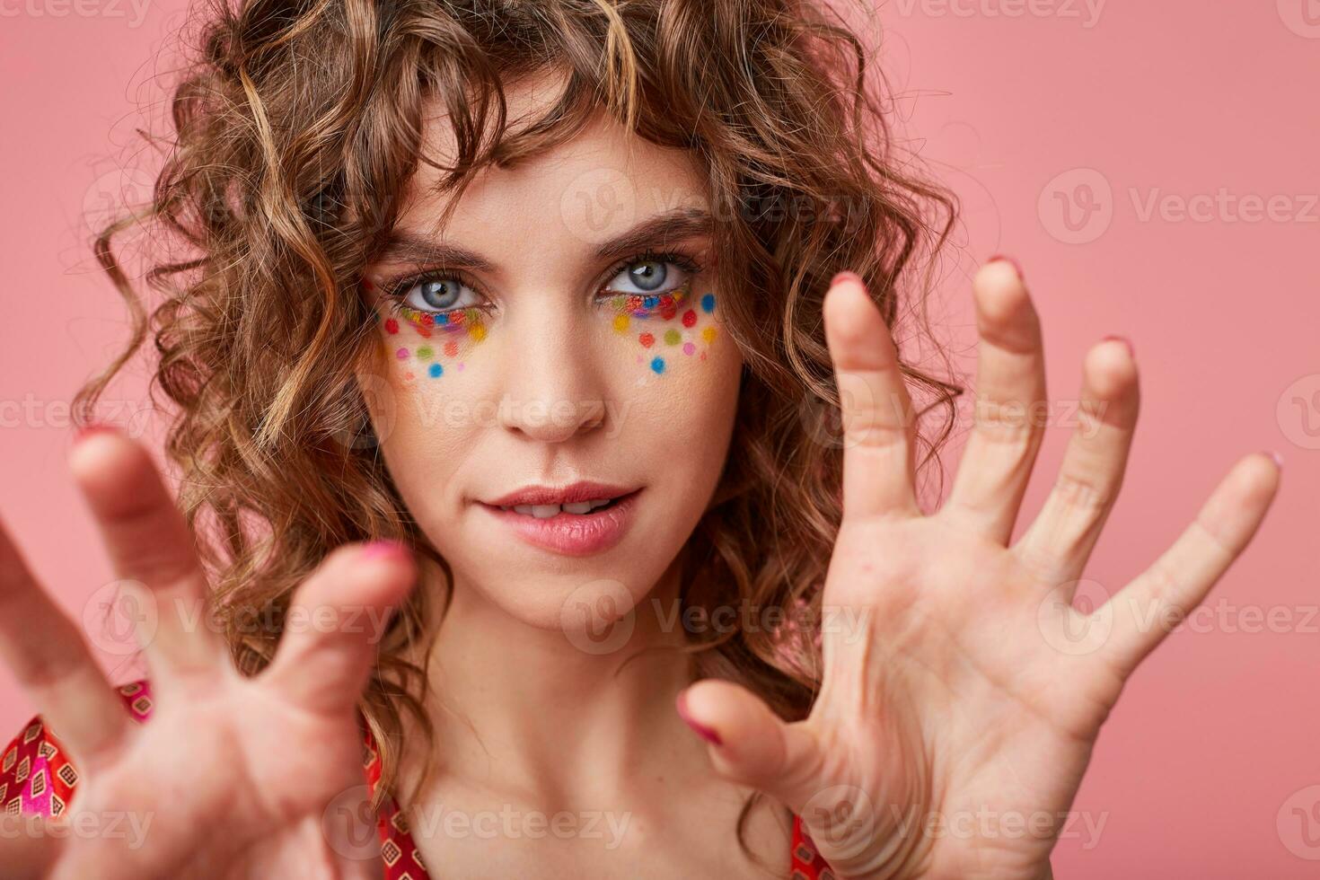 Close-up of flirty young blue-eyed woman with festive hairstyle and multicolored dots on her face biting underlip while posing over pink background with raised palms photo