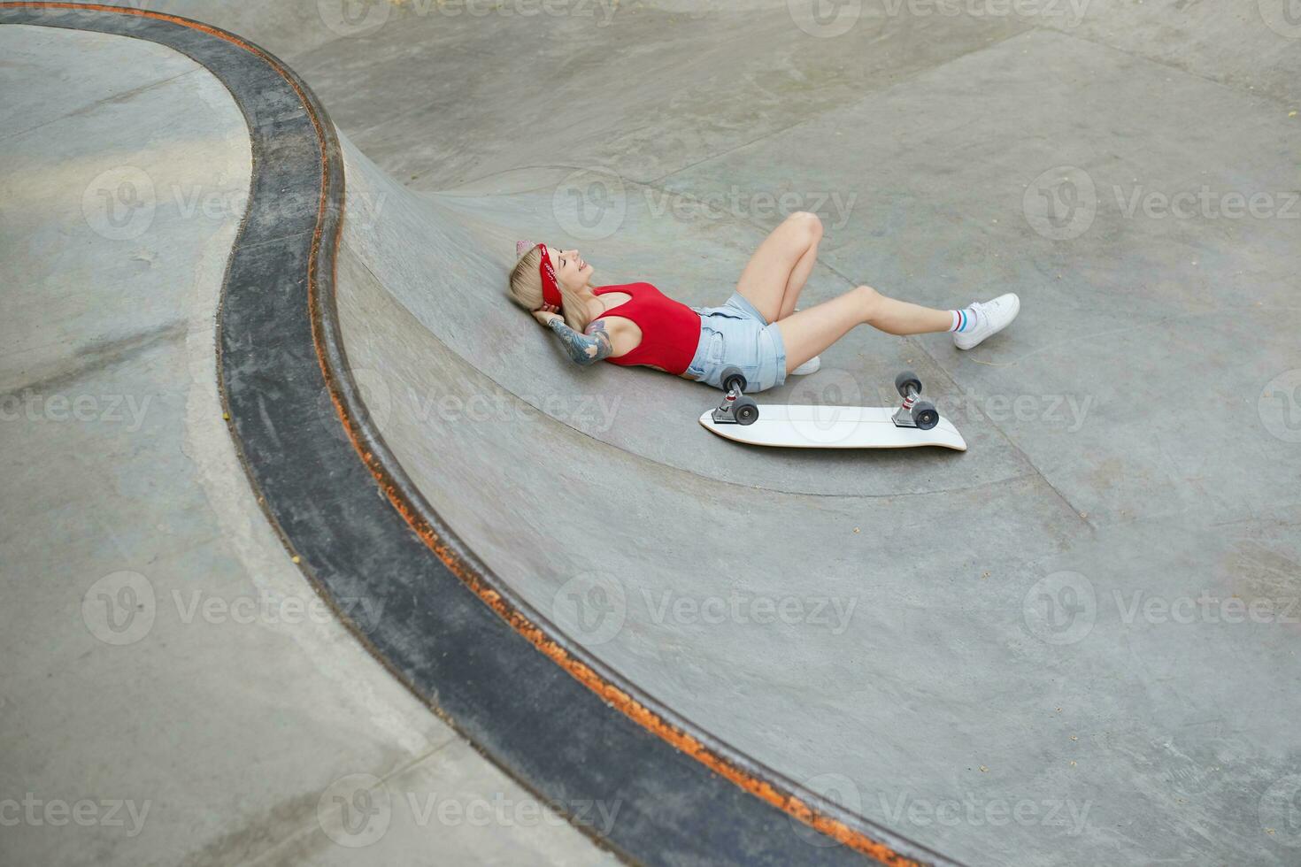 Outdoor shot of beautiful tattooed female in jeans shorts and red top relaxing over skate park on warm summer day, having break after skating photo