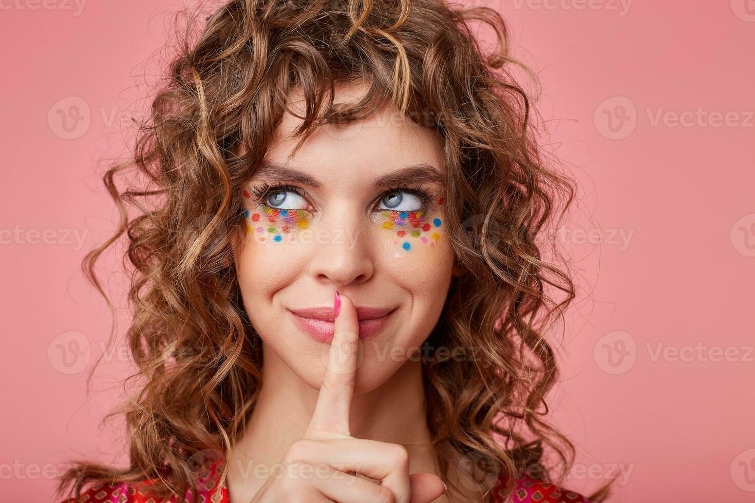 Cheerful young brunette curly lady with multicolored dots on her face looking aside with charming smile and keeping forefinger on her lips, isolated over pink background photo
