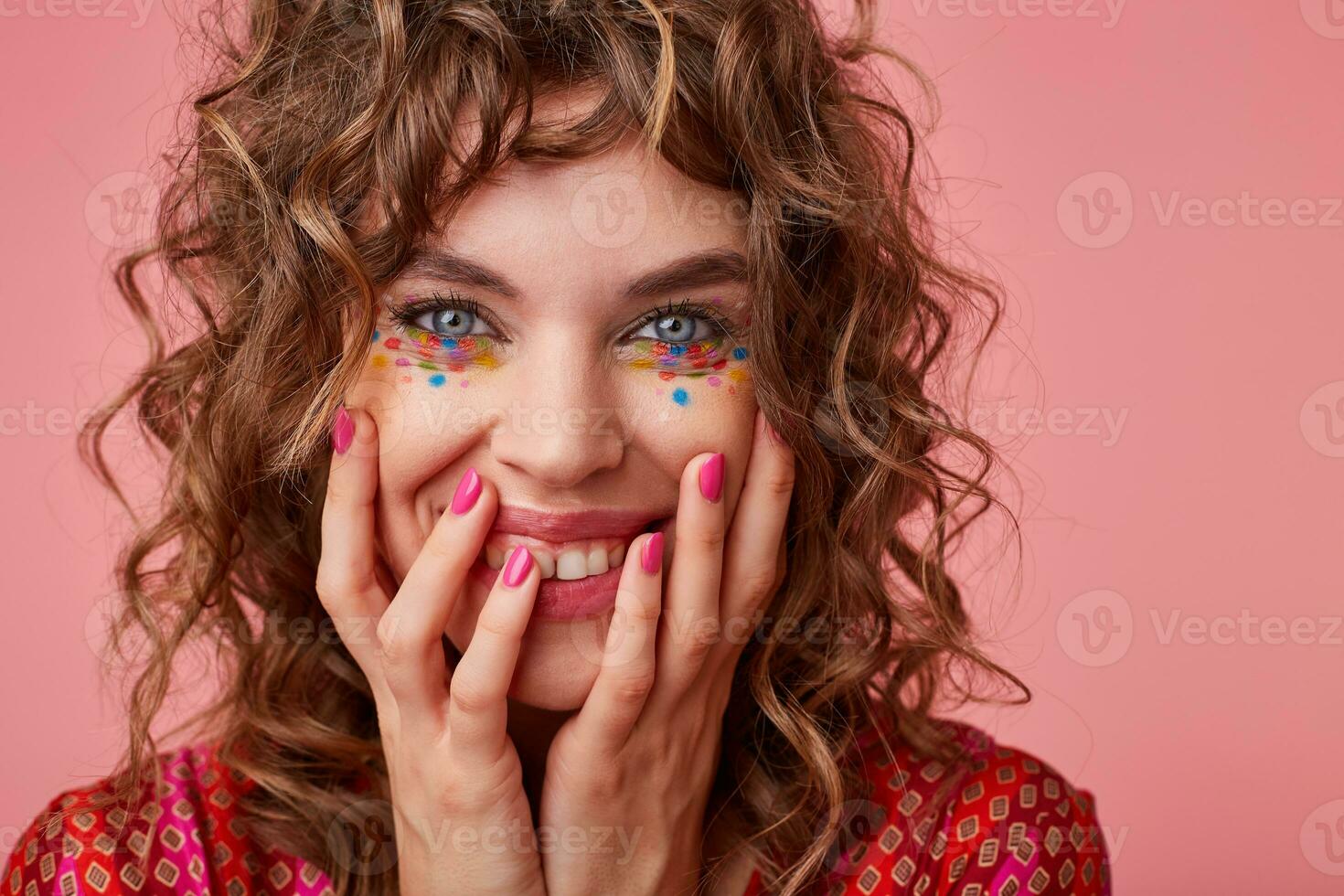 retrato de alegre joven Rizado mujer con festivo maquillaje participación su cara con manos y mirando a cámara con amplio sincero sonrisa, en pie terminado rosado antecedentes foto