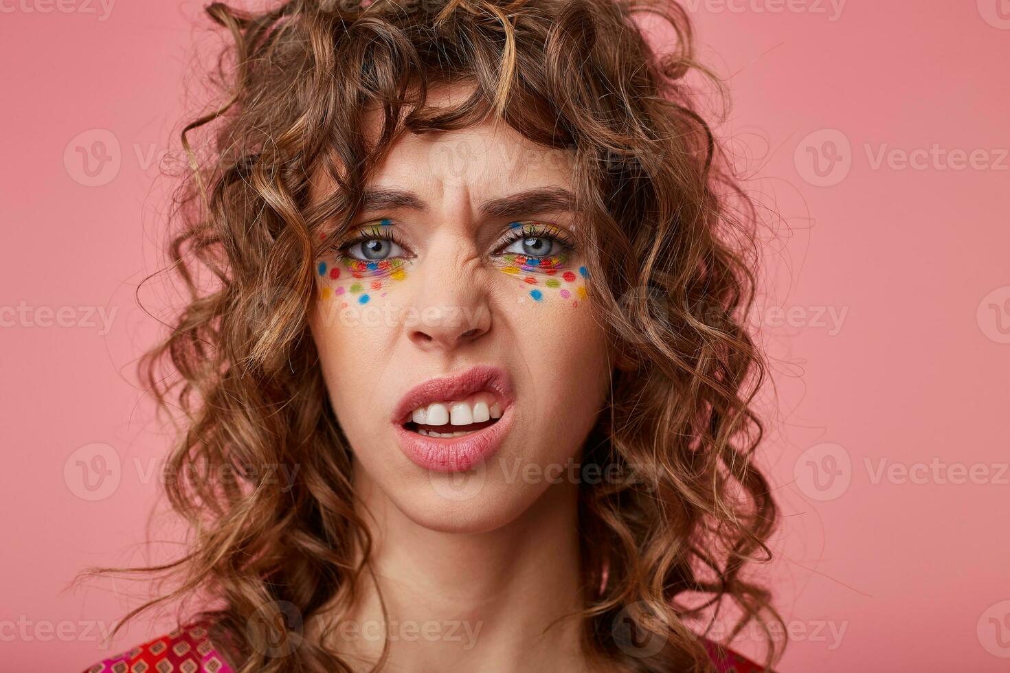 Close-up of dissatisfied curly blue-eyed lady with festive makeup posing over pink background, twisting her mouth and frowning eyebrows while looking at camera photo