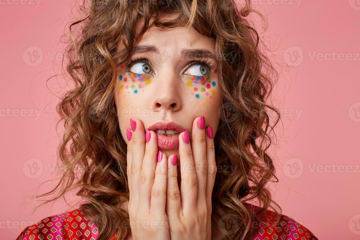 Frightened curly blue-eyed brunette female with multicolored dots on her face keeping hands on her face and looking scaredly aside, isolated over pink background photo